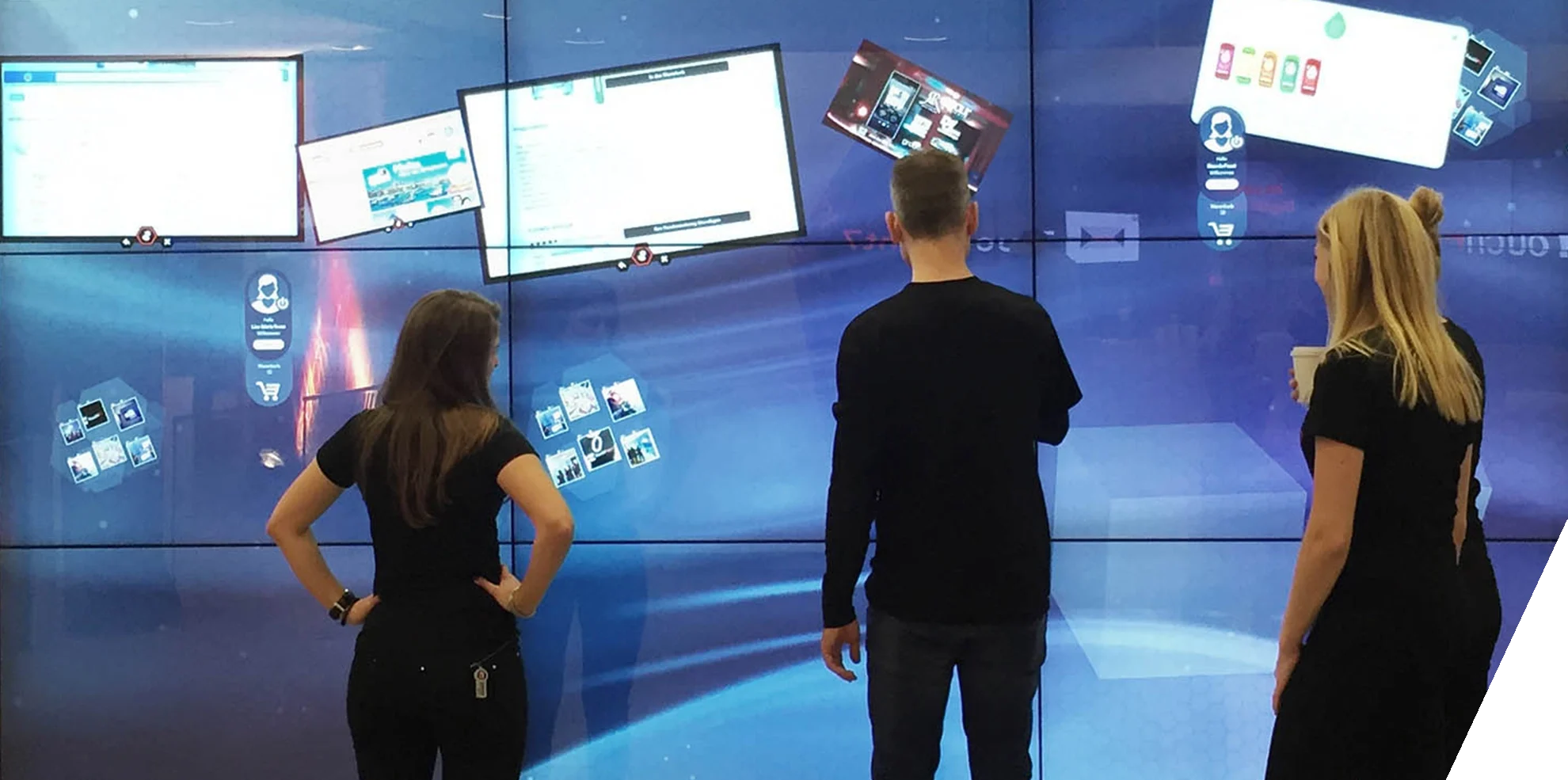 Drei Personen stehen vor einer Videowall. Eine Wand, die aus sechs Bildschirmen besteht und zusammen einen großen Screen darstellt.