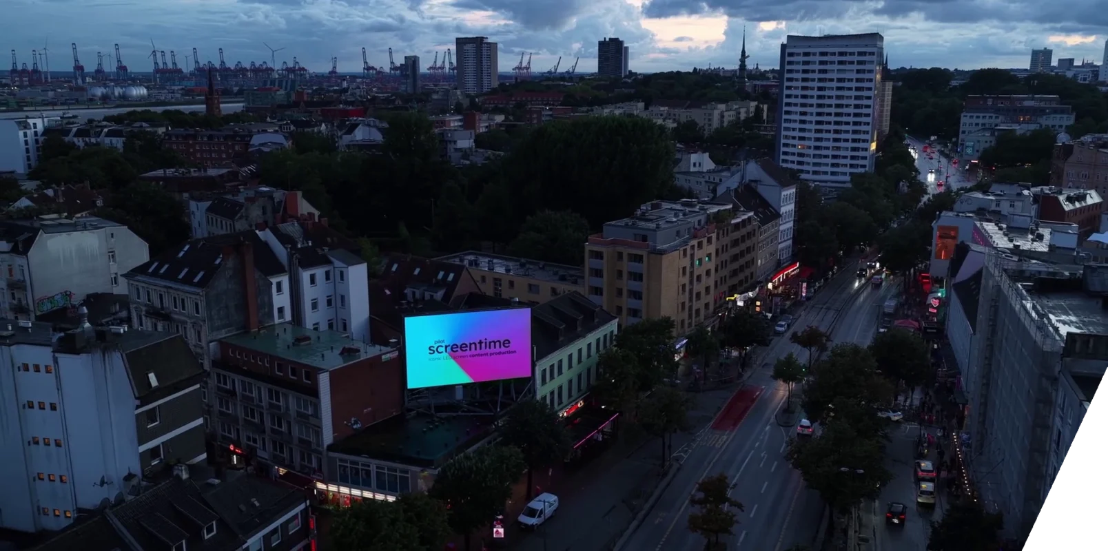 Ein Bild aus der Vogelperspektive zeigt die nächtliche Reeperbahn in Hamburg. Im Hintergrund sind Hafenkräne zu sehen. Zentral sieht man eine große LED Wall auf einem Hausdach.
